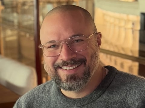 Smiling man with beard and glasses wearing a gray shirt, indoors with warm lighting